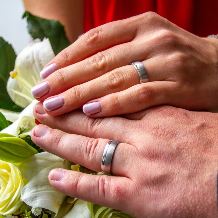 Hände mit Trauringen und Blumen, hinterlegt von einer roten Stoffoberfläche.