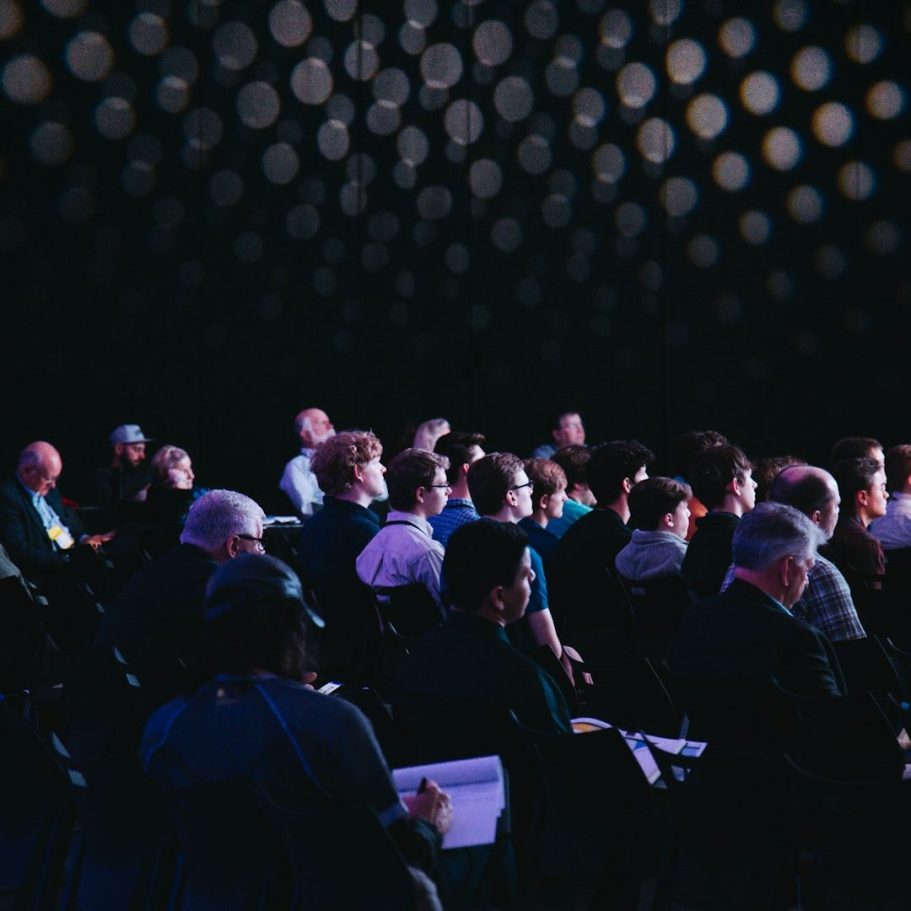 Veranstaltung mit Publikum, das gebannt auf die Bühne schaut, dunkler Raum mit Lichtpunkten.